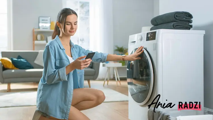 Kobieta kuca przed pralką sprawdzając telefon, poradnik jak czyścić zabrudzoną pralkę, czy da się idealnie wyczyścić pralkę