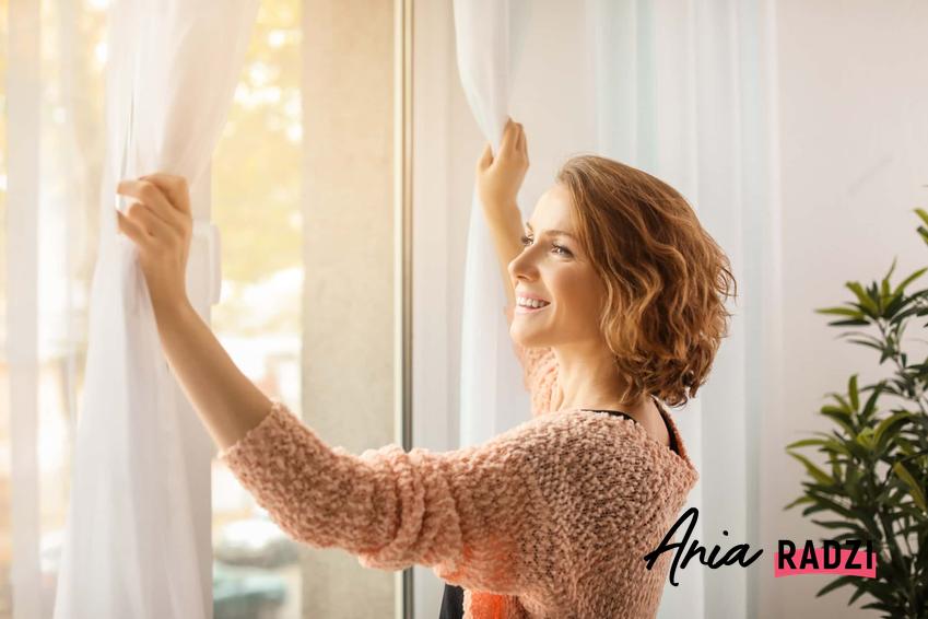 Kobieta odsłania firanki, kobieta wygląda przez okno, jak często prać firanki, jak prać firanki, żeby się nie zniszczyły
