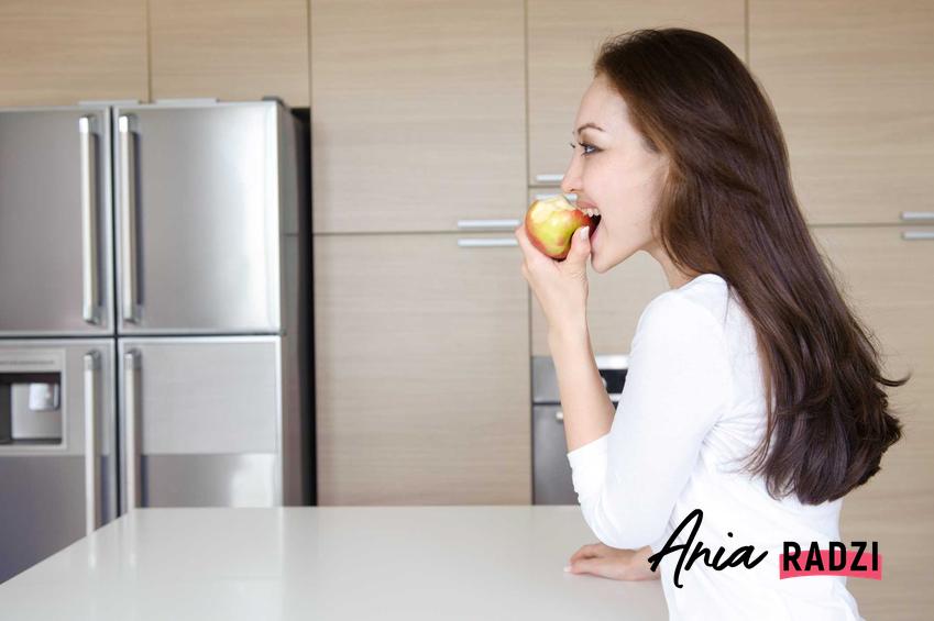 Kobieta gryząca jabłko obok amerykańskiej lodówki z podwójnymi drzwiami, czyli lodówki side by side krok po kroku
